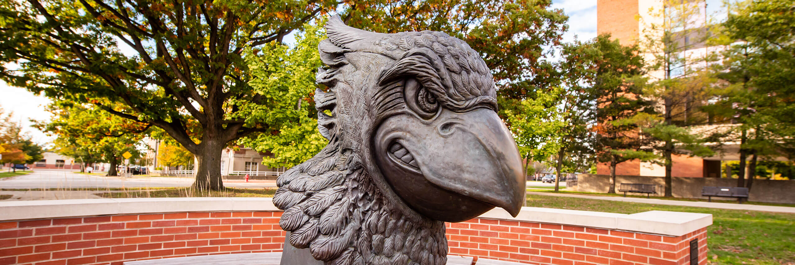 Bronze Raggie sculpture in front of trees and a brick building.